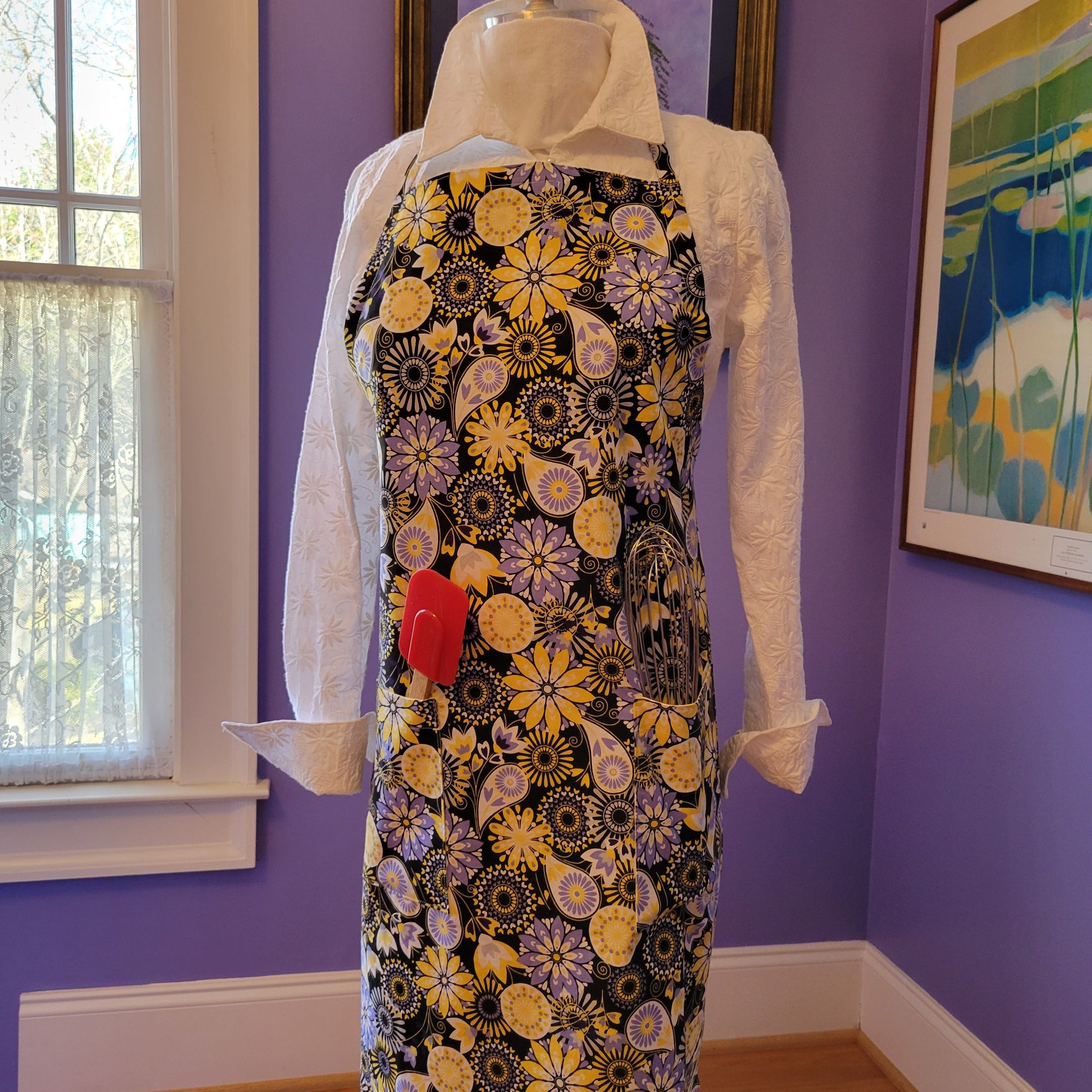 black, yellow, white and periwinkle flower fabric chef apron with red spatula tucked in 1 of the 2 pockets. Chef apron is displayed on a mannequin