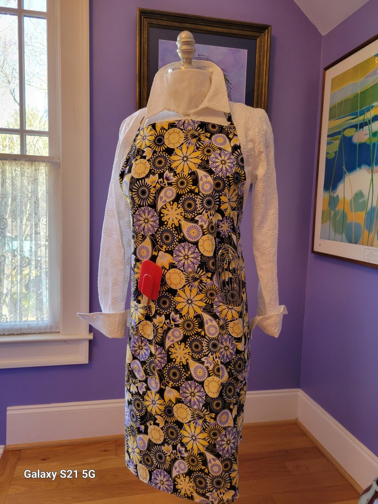 black, yellow, white and periwinkle flower fabric chef apron with red spatula tucked in 1 of the 2 pockets. Chef apron is displayed on a mannequin