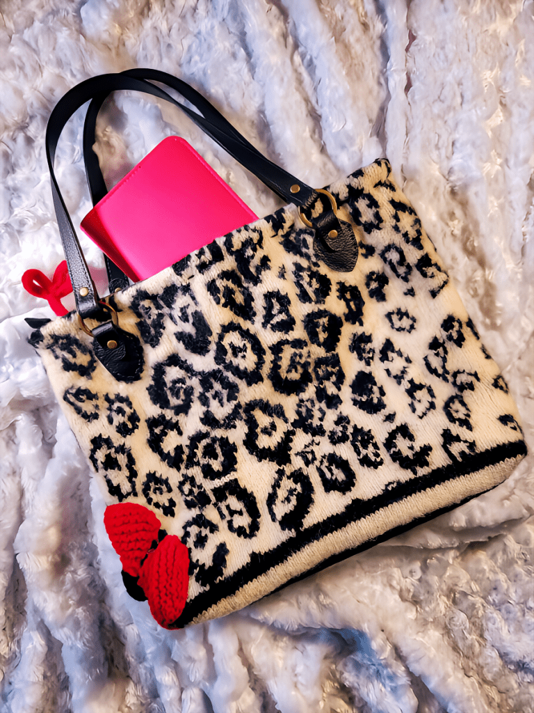 pic of upcycled handbag from discarded sweater with vegan leather straps and red flower petals on lower left side with pink book showing at top has fabric white background