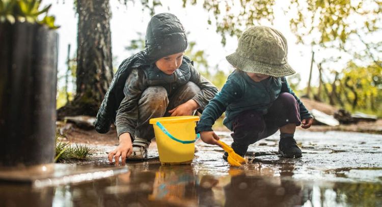 2 small kids playing with yellow bucket and shovel and dirt in wooded area